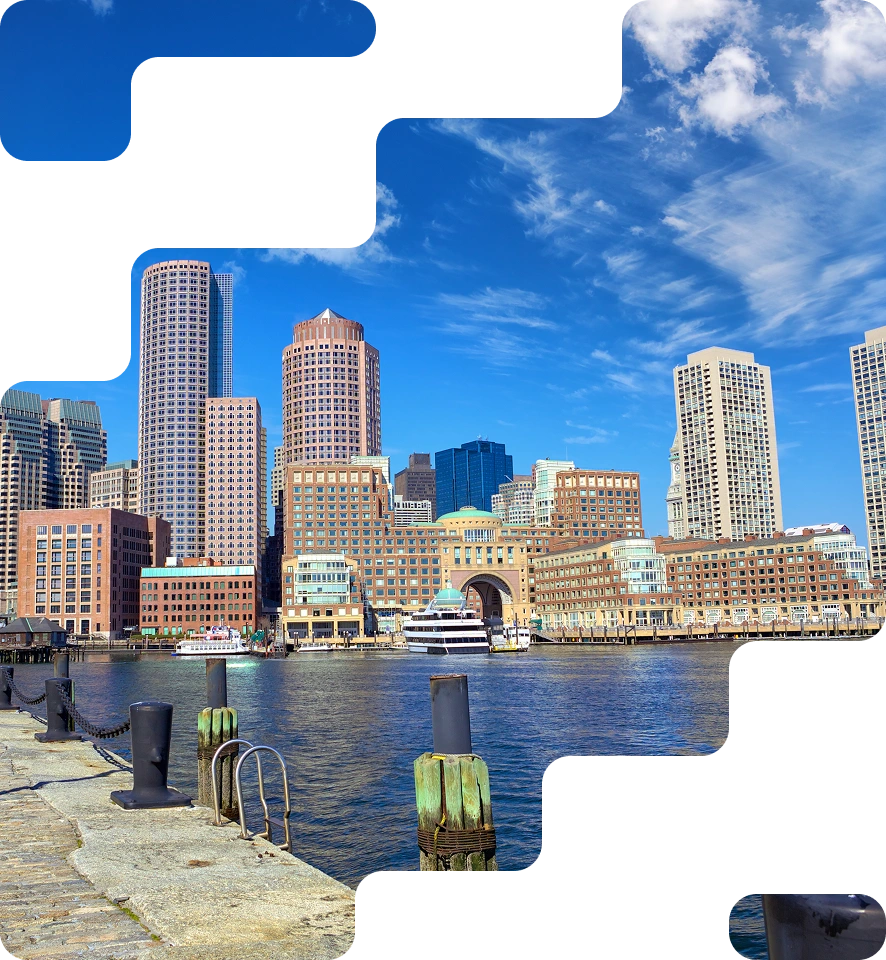 City skyline with tall modern buildings along a waterfront under a vibrant blue sky with wispy clouds. Foreground features a stone pier with mooring posts.