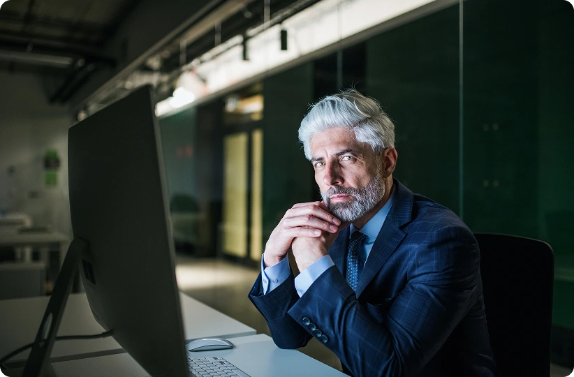 A man with gray hair and a beard, wearing a suit, sits thoughtfully at a desk, gazing at a large monitor. The office setting is dimly lit, conveying focus.