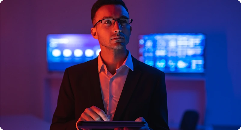 A man in a suit and glasses stands in a dimly lit room, looking pensive. He holds a tablet, with glowing blue screens behind him, suggesting a tech environment.