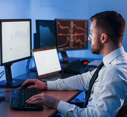 A man in a white shirt and tie works at a desk with multiple monitors displaying graphs and data in a dimly lit office, conveying focus and analysis.