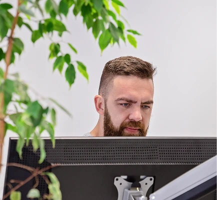 A man with a beard intensely focuses on a computer screen. A green plant is in the foreground, adding a touch of nature. The scene conveys concentration.