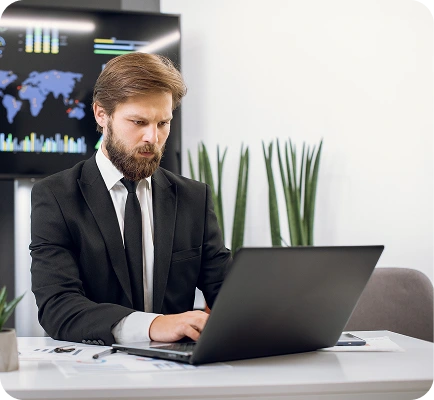 A man in a black suit works intently on a laptop at a modern office desk. Behind him, a world map with charts is displayed on a screen, adding a tech-savvy vibe.