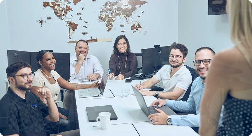 A diverse group of six people at a conference table, engaged in discussion with laptops; a world map decorates the wall, creating a collaborative and professional atmosphere.