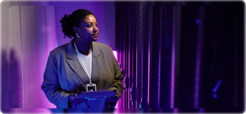 A person in a suit holds a tablet inside a dimly lit, purple-illuminated room, suggesting a data center or tech environment. The scene conveys focus and professionalism.